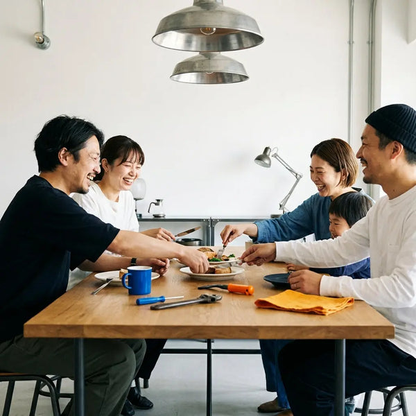 Gemütliche Szene bei babyparty ideen: enge Freunde/Familie am Esstisch, Hände greifen nach Tellern, warmes Licht, Holzoberfläche und dezente Metalldetails.