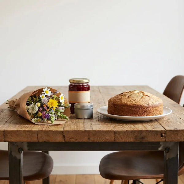 Nahaufnahme eines Holztischs mit typischen gastgeschenken deutschland Blumen Marmelade Metalldose und Vanillekuchen im warmen, leicht industriellen Esszimmer