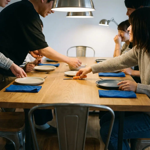 Kleine Familie an Esstisch mit Holzoberfläche und warmem Licht, Hände greifen nach Tellern, dezente Metalldetails und Akzente #0044CC/#ff9800 – was tun zum 1. hochzeitstag