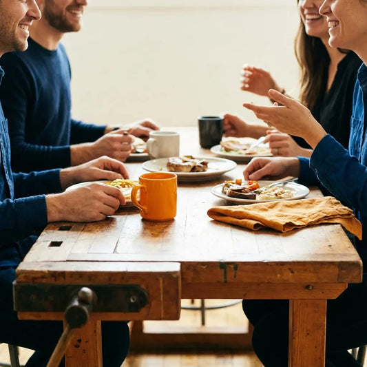 Wie viel schenkt man als Patenonkel? Kleine Familie am Esstisch mit warmem Licht, Holz- und Metalldetails, intime Zusammenkunft, Hände im Fokus, Gesichter nicht prominent.