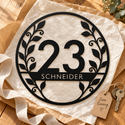Schwarzes botanisches Hausnummernschild 23 mit SCHNEIDER auf Geschenkpapier, neben Schlüsseln und dem Anhänger „Zum Einzug“.