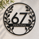 Schwarze botanische Hausnummer aus Metall mit 67 und MERTENS an einer hellen Putzwand.