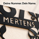 Nahaufnahme der ausgeschnittenen Schrift MERTENS in schwarzem Stahl mit den Hinweisen „Deine Nummer. Dein Name.“ und „2 mm Stahl“.